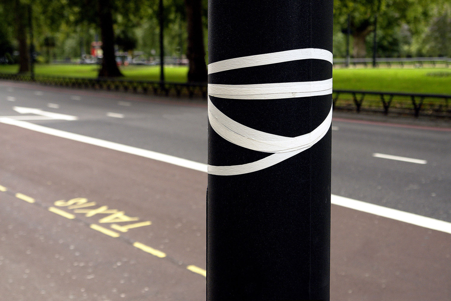 A black bollard wrapped in loops of white tape stands against a grey road with white lane markings and a yellow TAXIS legend painted on the tarmac